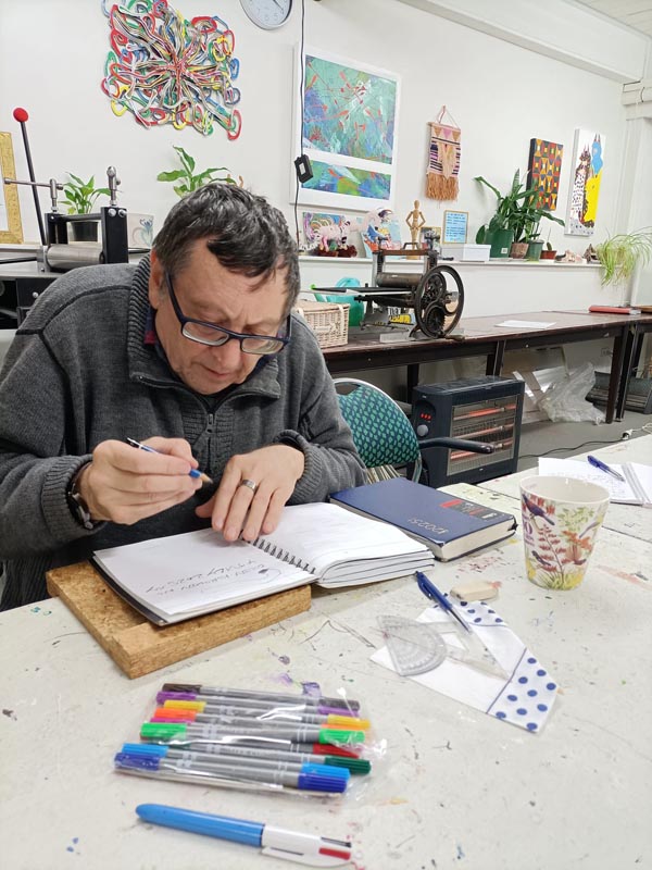 Artist Colin Korovin seat at a table, intent on sketching in one of his notebooks 